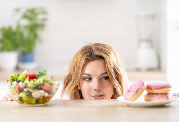 Beautiful young woman tempted having to make choice between fresh lettuce salad and doughnut in healthy unhealthy food, detox eating, calories and diet concept.