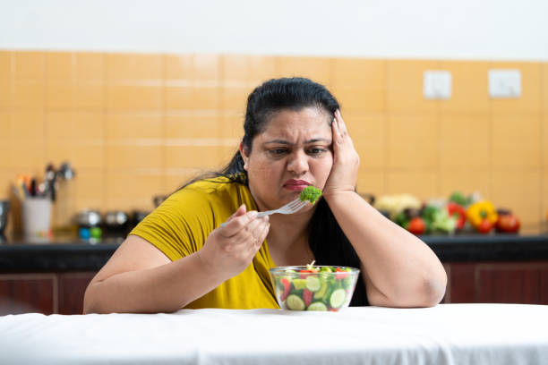 Sad Overweight fat indian woman holding fork and eating vegetable salad while sitting on a table in kitchen. Plus size female. Dieting struggle concept.