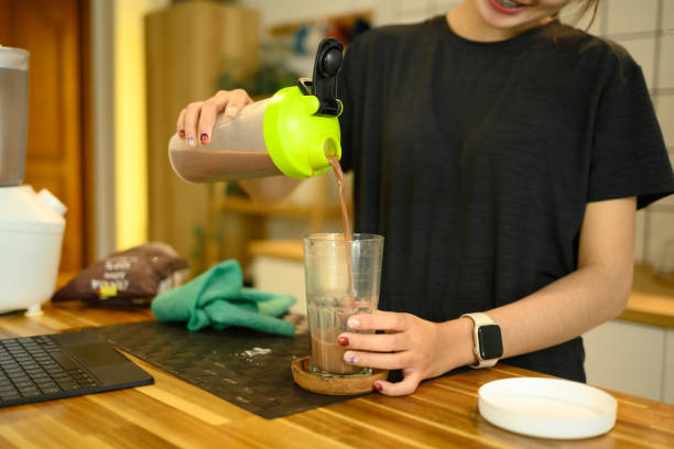 Smiling woman in sportswear pouring a freshly made chocolate protein shake into a glass.
